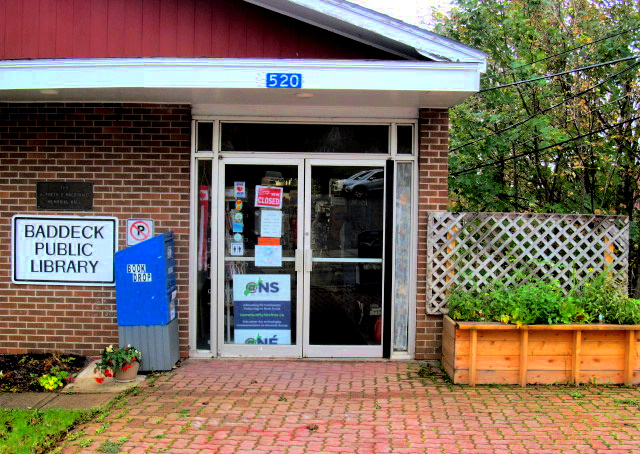 Exterior of Baddeck Library building showing Book returns bin to the left of the entrance doors and raised garden bed to the right of the entrance doors.