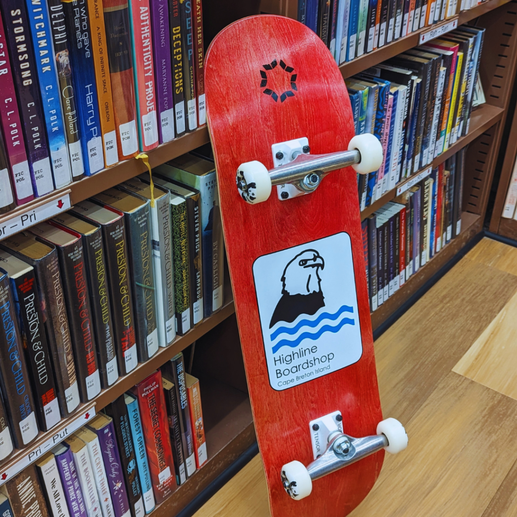 A skateboard leaning up against a shelf of library books. Underside of skateboard showing library logo and Highland Boardshop logo.  